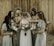 A group of women in elegant dresses surround a bride, gently placing their hands on her shoulders and back. Each woman holds a bouquet of flowers with pastel shades of pink, peach, and white. The setting appears rustic with a wooden backdrop, and two circular floral arrangements hang on the wall behind them.