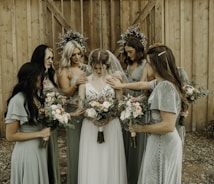 A group of women in elegant dresses surround a bride, gently placing their hands on her shoulders and back. Each woman holds a bouquet of flowers with pastel shades of pink, peach, and white. The setting appears rustic with a wooden backdrop, and two circular floral arrangements hang on the wall behind them.