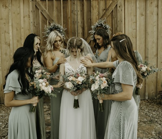 A group of women in elegant dresses surround a bride, gently placing their hands on her shoulders and back. Each woman holds a bouquet of flowers with pastel shades of pink, peach, and white. The setting appears rustic with a wooden backdrop, and two circular floral arrangements hang on the wall behind them.