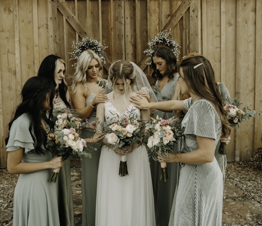 A group of women in elegant dresses surround a bride, gently placing their hands on her shoulders and back. Each woman holds a bouquet of flowers with pastel shades of pink, peach, and white. The setting appears rustic with a wooden backdrop, and two circular floral arrangements hang on the wall behind them.