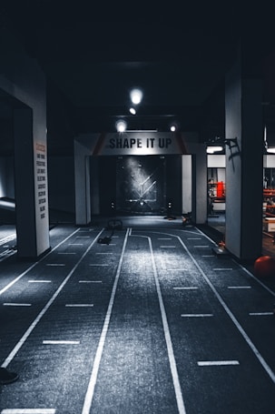 A dimly lit indoor gym with a central pathway marked with lane lines. The ceiling features multiple lights, and there are exercise equipment and motivational signage, including one that reads 'SHAPE IT UP'. The ambiance suggests a focus area for fitness activities.