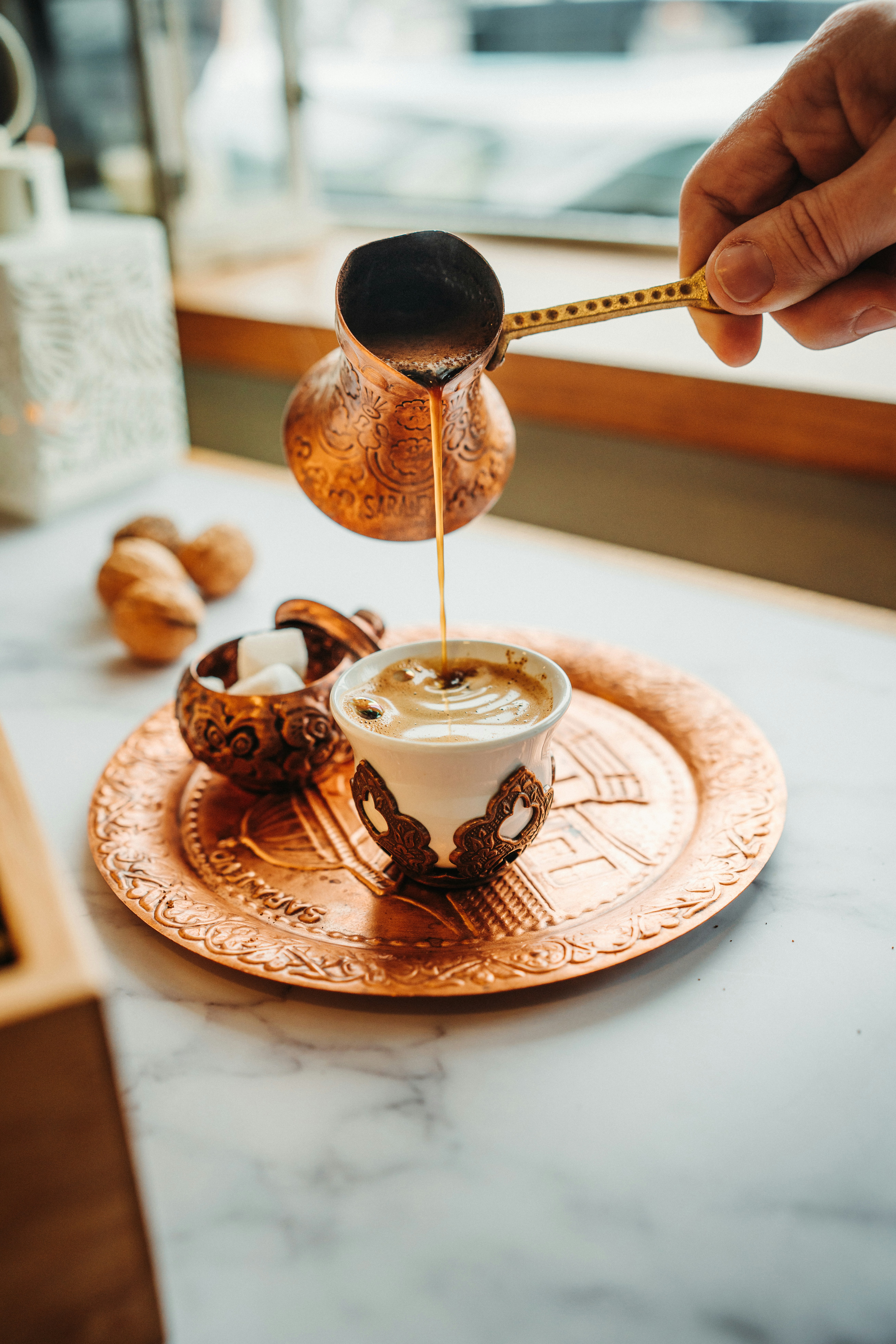A hand pouring coffee from a copper cezve into a porcelain cup on a decorated tray.