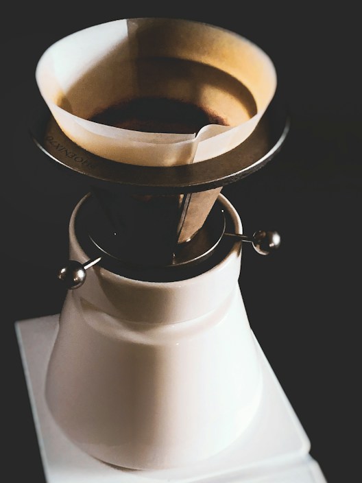 A coffee dripper resting on a ceramic carafe. The dripper contains a paper filter and ground coffee, with a metallic framework. The overall setup is placed against a dark background.