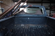 The interior of a pickup truck's cargo bed is visible, with a broken rear window through which wooden planks and debris have entered. An American flag is partially visible, draped under the planks. The scene suggests a recent impact or damage, possibly related to a storm or accident.