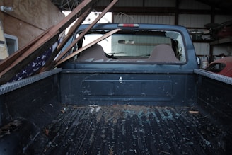 The interior of a pickup truck's cargo bed is visible, with a broken rear window through which wooden planks and debris have entered. An American flag is partially visible, draped under the planks. The scene suggests a recent impact or damage, possibly related to a storm or accident.