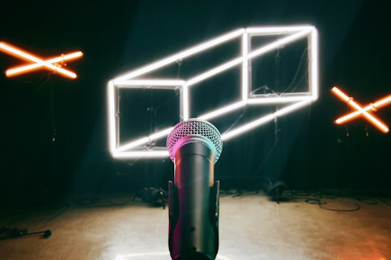 A microphone is positioned in the center foreground, with its grill clearly visible. Behind it, there are modern and geometric neon light structures, including white and orange LED lights forming shapes on a dark background.
