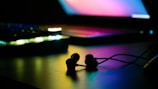 A pair of earbuds rests on a flat surface, subtly illuminated by colorful, ambient lighting. In the background, the blurred image of a laptop with a glowing screen adds depth to the scene, providing a modern and tech-focused atmosphere.