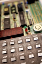 A close-up view of an electronic device featuring a keypad with various labeled buttons and a partially visible circuit board with a digital display. The buttons are gray and marked with labels like INS, SBR, DEL, and more. The circuit board in the background has multiple electronic components, including chips and connectors.
