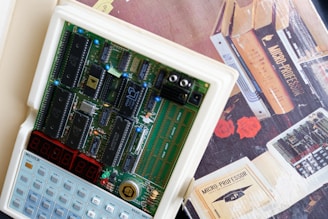 A vintage microcomputer kit with a visible circuit board containing various integrated circuits and components. Below the circuit board is a keypad with multiple buttons. The background includes a box with the text 'Micro-Professor' and illustrations of books and flowers.