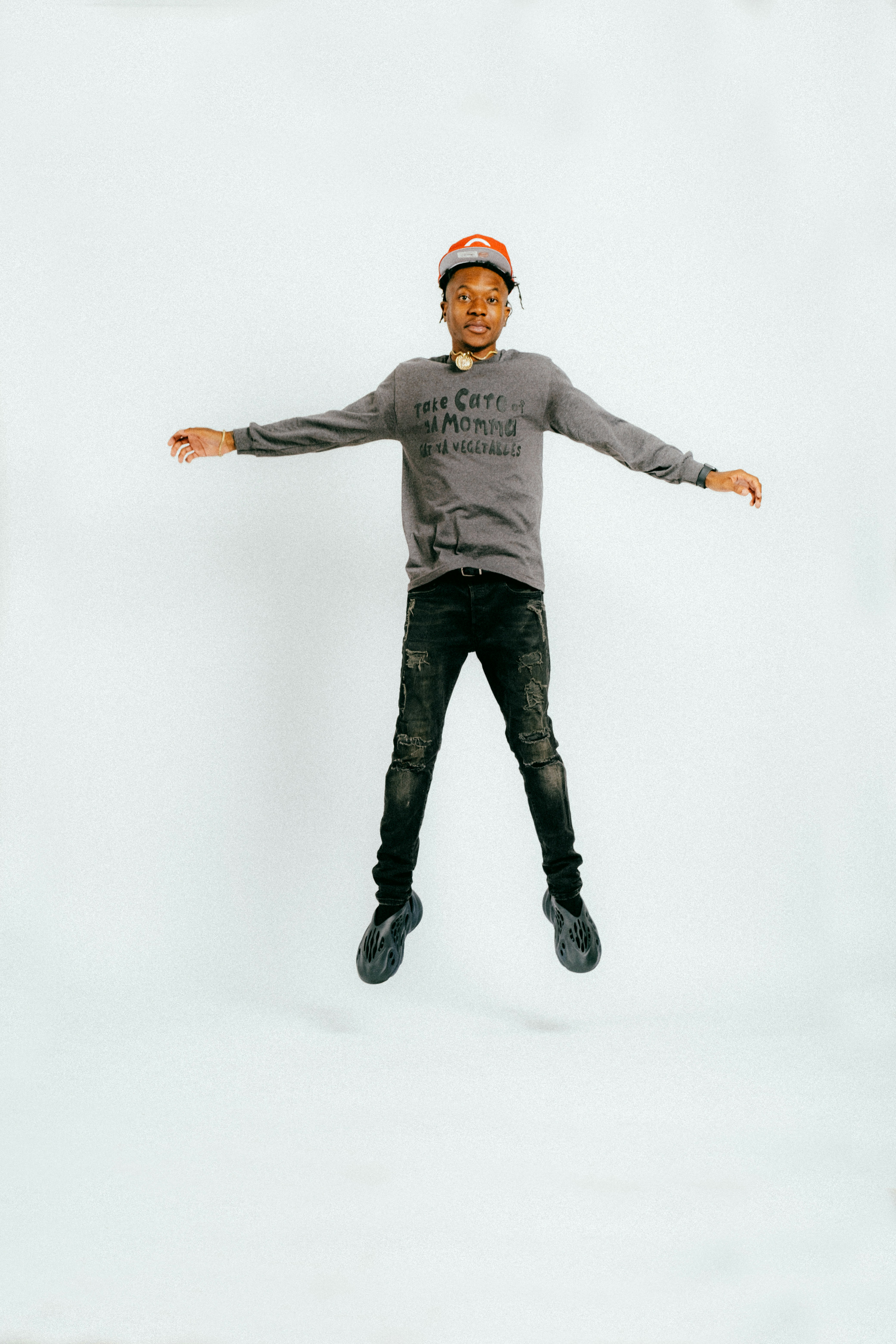 A young man in a cap and black jeans jumping with his arms spread against a white backdrop