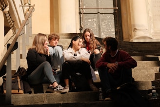 A group of five young people sit closely together on stone steps, engaged in conversation and enjoying each other's company. The setting appears to be outdoors, with sunlight casting warm tones over the scene. The group is dressed casually, with some carrying backpacks and water bottles nearby.