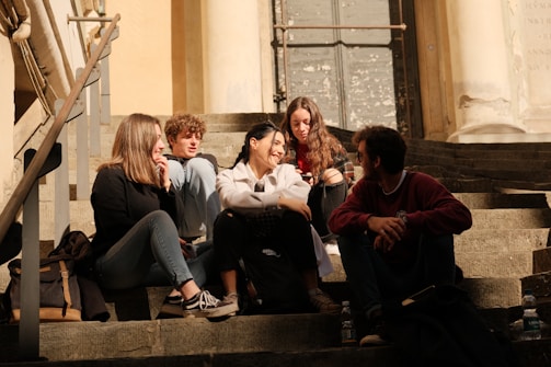 A group of five young people sit closely together on stone steps, engaged in conversation and enjoying each other's company. The setting appears to be outdoors, with sunlight casting warm tones over the scene. The group is dressed casually, with some carrying backpacks and water bottles nearby.