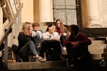 A group of five young people sit closely together on stone steps, engaged in conversation and enjoying each other's company. The setting appears to be outdoors, with sunlight casting warm tones over the scene. The group is dressed casually, with some carrying backpacks and water bottles nearby.