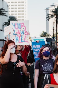 A group of people participate in a protest holding signs advocating for trans rights. One sign reads 'Your Laws Kill' with paint splashes, while another says 'Trans People Belong'. The participants are diverse in appearance, and some wear masks. The scene is set in an urban environment with palm trees and city buildings visible in the background.
