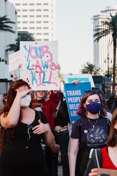 A group of people participate in a protest holding signs advocating for trans rights. One sign reads 'Your Laws Kill' with paint splashes, while another says 'Trans People Belong'. The participants are diverse in appearance, and some wear masks. The scene is set in an urban environment with palm trees and city buildings visible in the background.