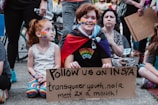 A group of people gathered, with one person holding a cardboard sign that reads 'Follow us on Insta: transqueer youth.nola meet 2x a month!' The person has rainbow face paint and is wearing a cape with a rainbow pattern and a shirt that says 'Protect Queer Kids.' A young child beside them with rainbow face paint is also present. Other individuals in the background are standing or sitting, and some hold signs.