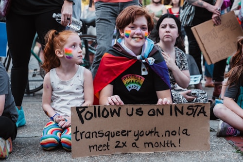 A group of people gathered, with one person holding a cardboard sign that reads 'Follow us on Insta: transqueer youth.nola meet 2x a month!' The person has rainbow face paint and is wearing a cape with a rainbow pattern and a shirt that says 'Protect Queer Kids.' A young child beside them with rainbow face paint is also present. Other individuals in the background are standing or sitting, and some hold signs.