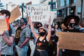 A group of people are participating in a protest on a city street, holding various signs advocating for LGBTQ+ rights. The individuals are wearing masks, and the mood appears assertive and determined. Buildings and palm trees can be seen in the background.