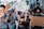 A group of people are participating in a protest on a city street, holding various signs advocating for LGBTQ+ rights. The individuals are wearing masks, and the mood appears assertive and determined. Buildings and palm trees can be seen in the background.