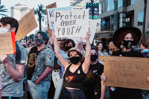 A group of people are participating in a protest on a city street, holding various signs advocating for LGBTQ+ rights. The individuals are wearing masks, and the mood appears assertive and determined. Buildings and palm trees can be seen in the background.