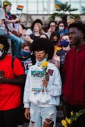 A group of diverse young people, some wearing face masks, gather outdoors holding protest signs and flowers. The scene includes a person in the foreground with yellow flowers and a flyer that discusses activism. In the background, rainbow flags are visible along with other individuals.