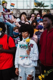 A group of diverse young people, some wearing face masks, gather outdoors holding protest signs and flowers. The scene includes a person in the foreground with yellow flowers and a flyer that discusses activism. In the background, rainbow flags are visible along with other individuals.