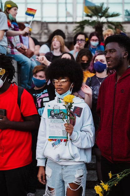 A group of diverse young people, some wearing face masks, gather outdoors holding protest signs and flowers. The scene includes a person in the foreground with yellow flowers and a flyer that discusses activism. In the background, rainbow flags are visible along with other individuals.