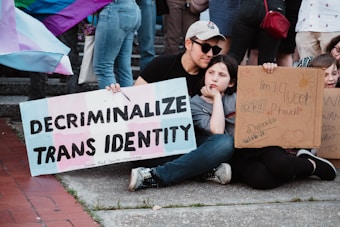 A group of individuals is sitting on the ground holding signs advocating for transgender rights. One sign prominently displays the message 'Decriminalize Trans Identity.' Another sign contains handwritten messages expressing queer pride and support. There are various pride flags in the background, and the setting appears to be outdoors on a paved area.