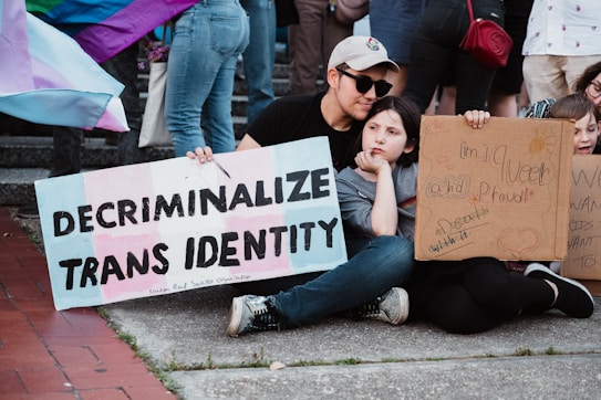 A group of individuals is sitting on the ground holding signs advocating for transgender rights. One sign prominently displays the message 'Decriminalize Trans Identity.' Another sign contains handwritten messages expressing queer pride and support. There are various pride flags in the background, and the setting appears to be outdoors on a paved area.