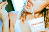 A person in a white tank top is eating noodles from a cup using a fork. The scene focuses on the mouth and hand holding the cup, which is branded with 'Maruchan Instant Lunch'. The lighting gives a warm, slightly overexposed appearance.