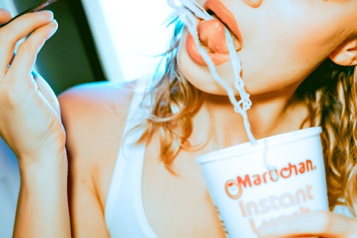 A person in a white tank top is eating noodles from a cup using a fork. The scene focuses on the mouth and hand holding the cup, which is branded with 'Maruchan Instant Lunch'. The lighting gives a warm, slightly overexposed appearance.