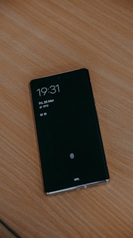 A smartphone with a black screen displaying the time as 19:31, the date as Friday, 25 March, and a temperature of 19°C. The phone is placed on a wooden surface and shows icons for notifications at the bottom.