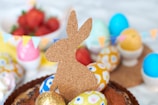 A corkboard silhouette of a rabbit is surrounded by colorful, decorated Easter eggs and vibrant strawberries in the background. Small triangular flags are strung across, adding to the festive atmosphere. The scene is set with a focus on spring and celebration.