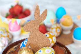 A corkboard silhouette of a rabbit is surrounded by colorful, decorated Easter eggs and vibrant strawberries in the background. Small triangular flags are strung across, adding to the festive atmosphere. The scene is set with a focus on spring and celebration.