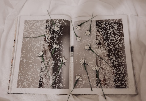 A book is open to a page with a monochrome photograph covered in white petals. Several white flowers with green stems are placed randomly across the pages, creating a layered, artistic effect. The background features a soft, white fabric, contributing to a delicate and serene atmosphere.