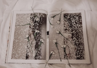 A book is open to a page with a monochrome photograph covered in white petals. Several white flowers with green stems are placed randomly across the pages, creating a layered, artistic effect. The background features a soft, white fabric, contributing to a delicate and serene atmosphere.