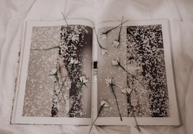 A book is open to a page with a monochrome photograph covered in white petals. Several white flowers with green stems are placed randomly across the pages, creating a layered, artistic effect. The background features a soft, white fabric, contributing to a delicate and serene atmosphere.