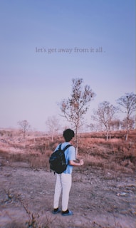 A person stands in a dry, sparsely wooded landscape with fields of brown grass and trees with minimal foliage. The sky is clear and blue. The person is wearing a light blue shirt, white pants, and a black backpack, facing towards the trees. Text overlay reads 'let's get away from it all.'