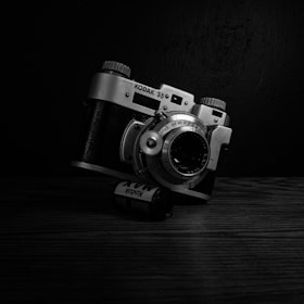 A vintage Kodak 35mm film camera is positioned at an angle on a wooden surface. A film roll, labeled Kodak, is placed in front of the camera. The image is in black and white, providing a nostalgic feel with high contrast against a dark background.