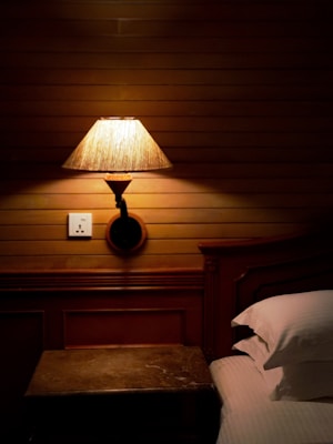 A warmly lit wooden paneled wall with a vintage bedside lamp casting a soft glow. A wooden bed with white pillows partially visible, accompanied by a small wooden nightstand. An electrical outlet is positioned near the lamp.