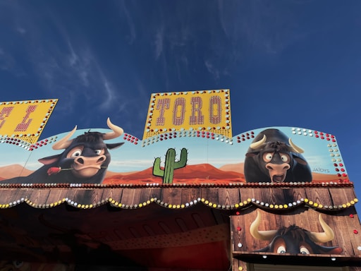 A colorful amusement ride decoration featuring two cartoon bulls with large horns, one holding a rose in its mouth. A cactus is depicted between them against a backdrop of desert hills and a clear blue sky. The signage reads 'TORO' in bright yellow letters, decorated with small lights.