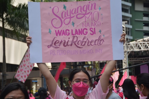 A person in a pink shirt and pink face mask holds up a rectangular pink sign featuring text in both capital and cursive fonts. Around them are other people, some wearing pink, and in the background, there is a stage structure along with a few palm trees and a building.