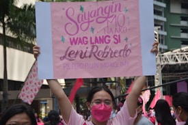 A person in a pink shirt and pink face mask holds up a rectangular pink sign featuring text in both capital and cursive fonts. Around them are other people, some wearing pink, and in the background, there is a stage structure along with a few palm trees and a building.