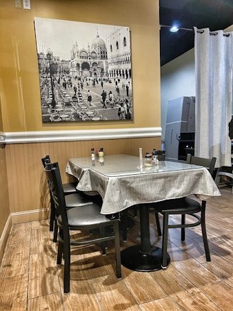 A cozy dining area with a table set for four, covered with a light gray tablecloth and accompanied by black chairs. The wall features a large black and white photograph depicting a historic European square with grand architecture and people walking by. The flooring is wood-like, complementing the warm, neutral tones of the room.