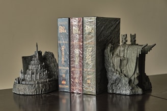 The image features a set of three DVD box sets of 'The Lord of the Rings' placed between two detailed, gray stone bookends. The bookends depict large, intricate structures with one resembling a fortress or city on a hill, and the other featuring two towering human-like figures in robes with outstretched arms. The DVDs are encased in plastic wrap, featuring the titles 'The Fellowship of the Ring,' 'The Two Towers,' and 'The Return of the King.'