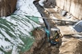 A construction site with a small excavator operated by a person clearing snow and dirt along a trench. The trench is lined with a tarp and is bordered by large pipes and wooden planks. Significant patches of snow cover the ground, particularly on the sides of the trench.