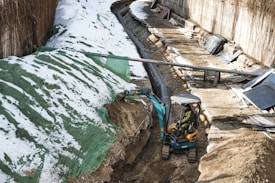 A construction site with a small excavator operated by a person clearing snow and dirt along a trench. The trench is lined with a tarp and is bordered by large pipes and wooden planks. Significant patches of snow cover the ground, particularly on the sides of the trench.