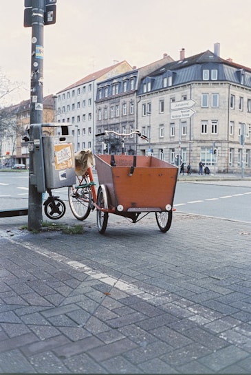 A red cargo bicycle with a wooden compartment is parked on a paved street corner next to a pole covered with stickers and a trash bin. In the background, there are multi-story residential and commercial buildings with a traditional architectural style.