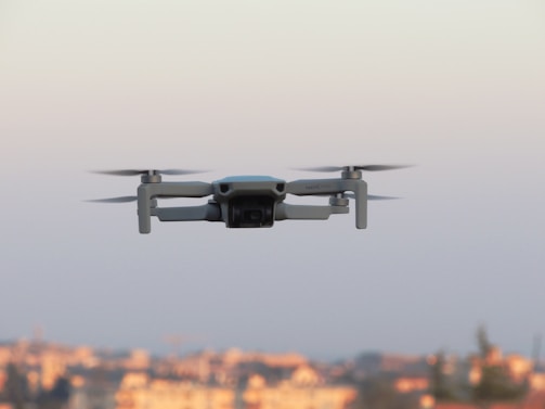 A small quadcopter drone hovering in the sky with a blurred background that suggests a distant cityscape.