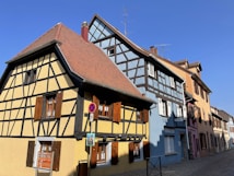 A charming street featuring traditional half-timbered houses with wooden accents and pastel-colored facades, including shades of yellow and blue. The roofs are steep and covered with reddish-brown tiles. The cobblestone street is empty, and the sky is clear, indicating a sunny day. Visible are some signs attached to the buildings and streets, adding character to the scene.